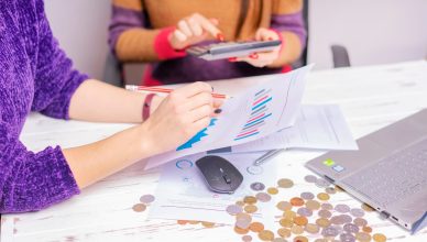 Two women analyzing financial charts with a laptop and coins on desk in an office setting. — 4 amazing Forex career for the r