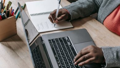 Crop unrecognizable African American female in formal wear writing notes in notepad while working on laptop — A demo account