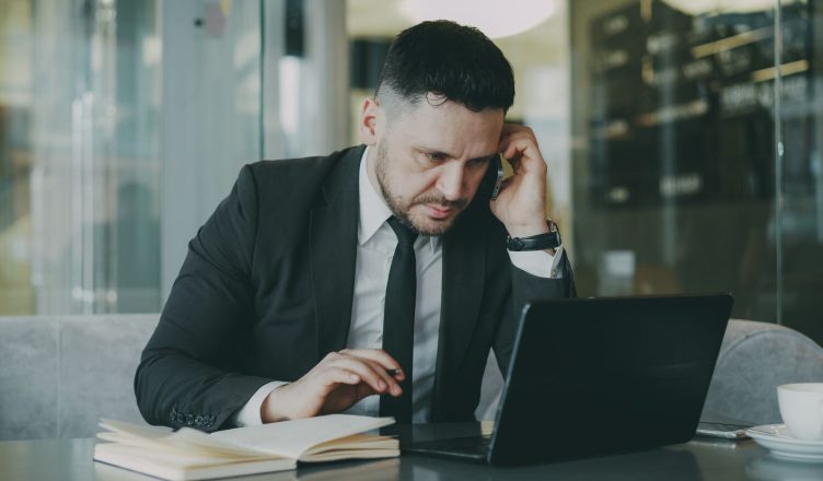 Focused businessman in suit multitasking on phone and laptop in modern office. — All you need to know about Dropshipping and