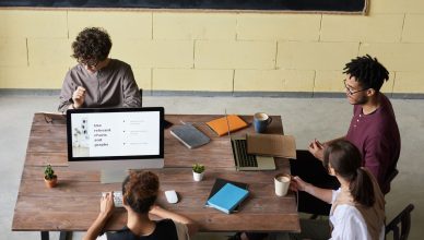 Four diverse colleagues engaged in a team meeting, working on laptops with coffee and notes, fostering collaboration. — Benef