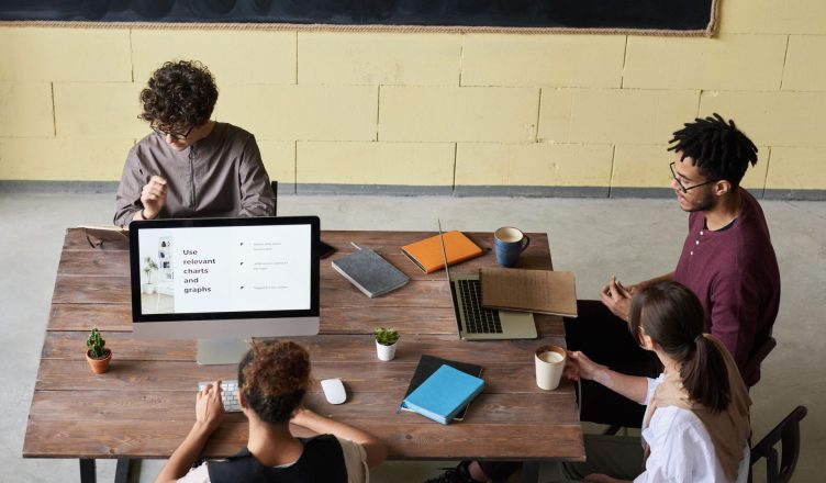 Four diverse colleagues engaged in a team meeting, working on laptops with coffee and notes, fostering collaboration. — Benef