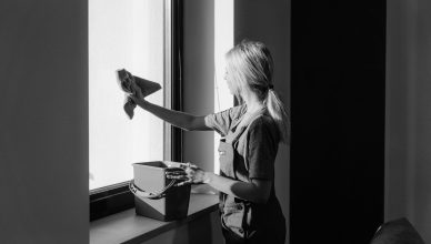Black and white image of a woman cleaning a window with a cloth and bucket indoors. — Growing Demand For Green Cleaning Solut