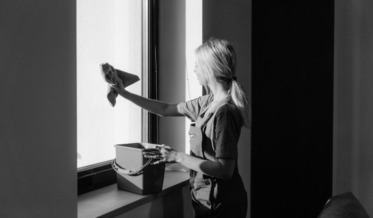 Black and white image of a woman cleaning a window with a cloth and bucket indoors. — Growing Demand For Green Cleaning Solut