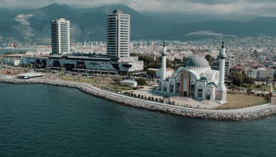 Aerial photograph showcasing İskenderun's mosque and cityscape against the mountainous backdrop on a sunny day. — How can you