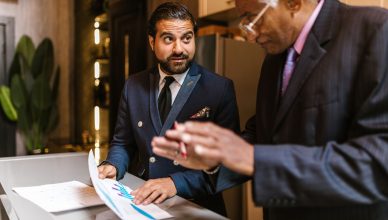 Two men in suits discuss financial documents with graphs indoors. — How to Buy a New Rental Property