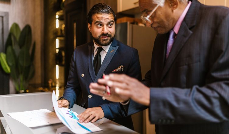Two men in suits discuss financial documents with graphs indoors. — How to Buy a New Rental Property