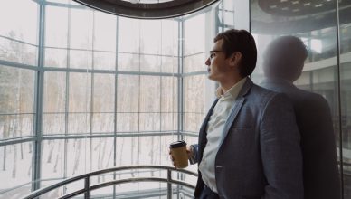 Businessman in blue suit with coffee cup looking out a large window, reflecting on a winter landscape. — How to Diversify You