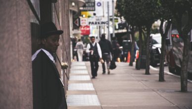 A man in a suit and hat leans against a city building, observing pedestrians. — How to Find the Best Life Insurance Company