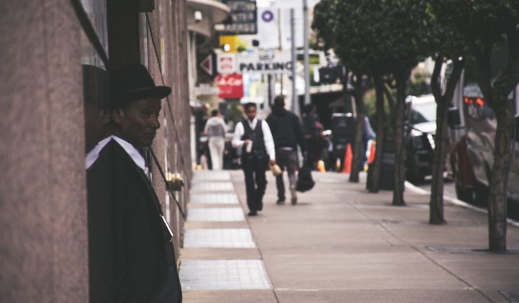A man in a suit and hat leans against a city building, observing pedestrians. — How to Find the Best Life Insurance Company