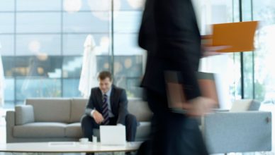 Businessman sitting in modern office lounge using a laptop while colleague walks past, creating dynamic blur. — How to Maximi