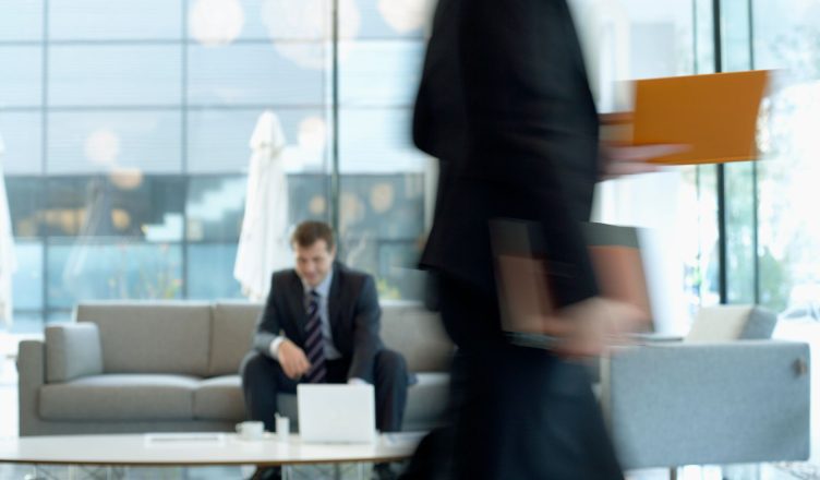 Businessman sitting in modern office lounge using a laptop while colleague walks past, creating dynamic blur. — How to Maximi