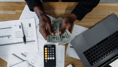 Person counting US dollars, using a calculator and laptop, with financial documents on a wooden desk. — Pros and Cons of VA M
