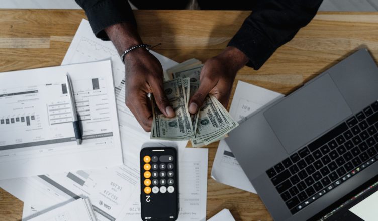 Person counting US dollars, using a calculator and laptop, with financial documents on a wooden desk. — Pros and Cons of VA M