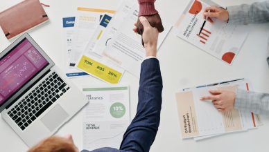Business professionals engaging in a handshake over a desk filled with financial documents and a laptop. — Look and F