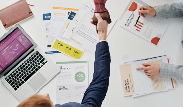 Business professionals engaging in a handshake over a desk filled with financial documents and a laptop. — Look and F