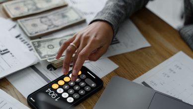Close-up of a person using a smartphone calculator amid money and financial documents on a wooden table. — What to Expect Fro