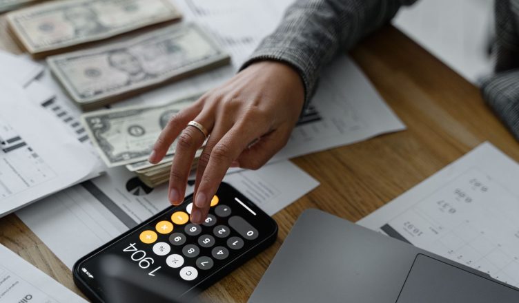 Close-up of a person using a smartphone calculator amid money and financial documents on a wooden table. — What to Expect Fro