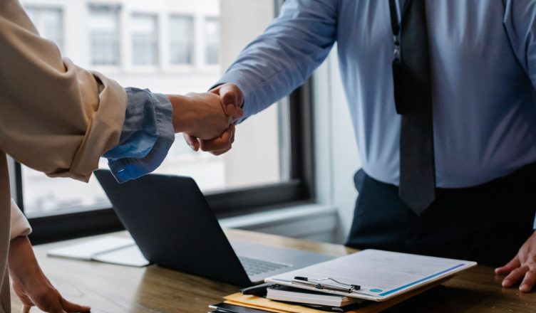 Crop unrecognizable coworkers in formal wear standing at table with laptop and documents while greeting each other before mee