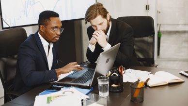 Two businessmen engaged in a strategy meeting with laptops and charts. — Working on landscaping insurance, and who is it righ